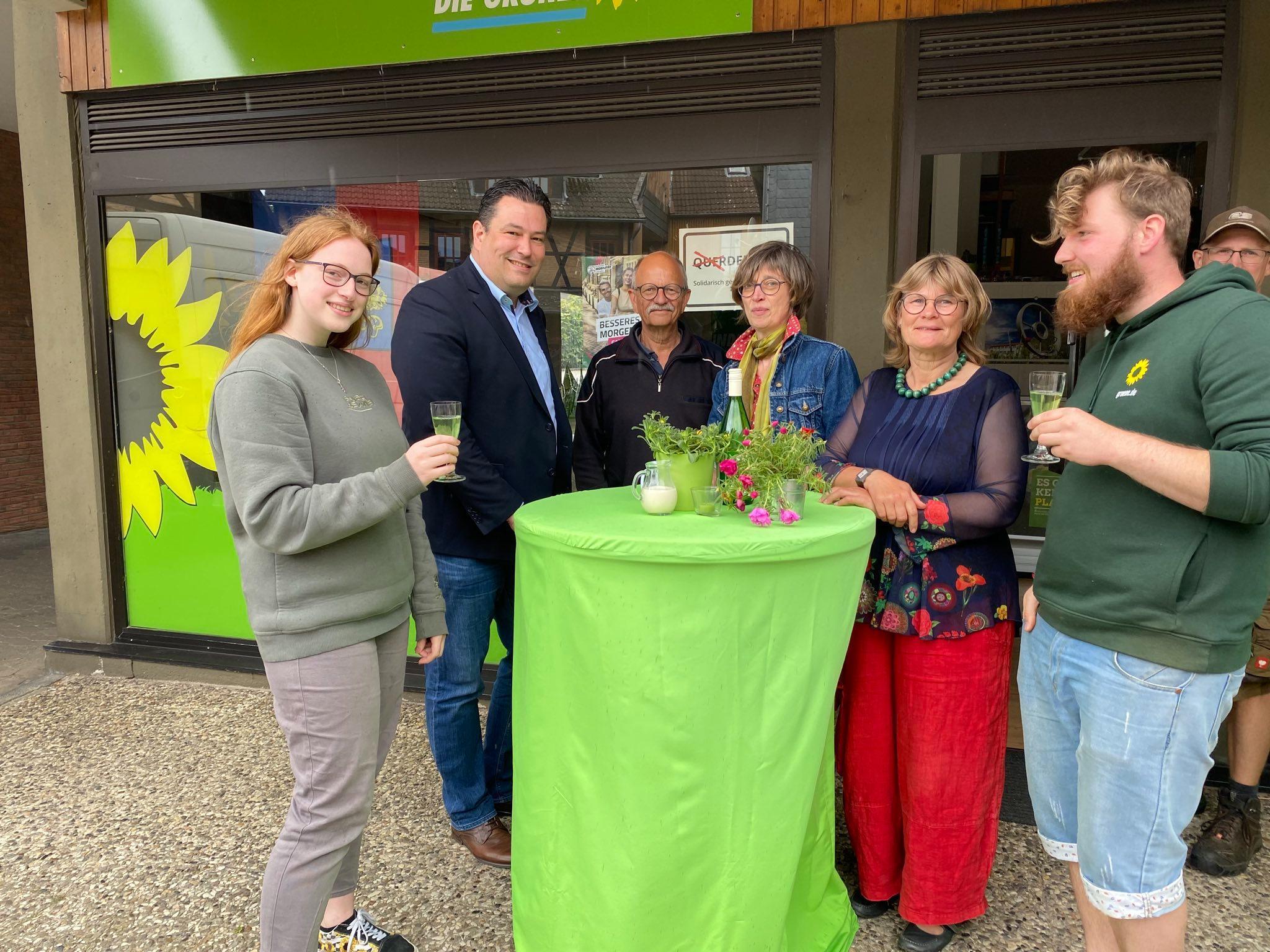 Mitglieder der Grünen und der Grünen Jugend zusammen mit Landrat Heilmann vor der Grünen Geschäftsstelle in Gifhorn. Sie trinken grüne Maibowle und stehen um einen grünen Tisch.