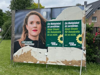 Ein beschädigtes Großplakat der Grünen mit zwei Zusatzplakaten darüber mit dem Text "Ein Wahlplakat ist zerstörbar. Unser Einsatz für Demokratie ist es nicht. gruene.de, Machen was zählt."