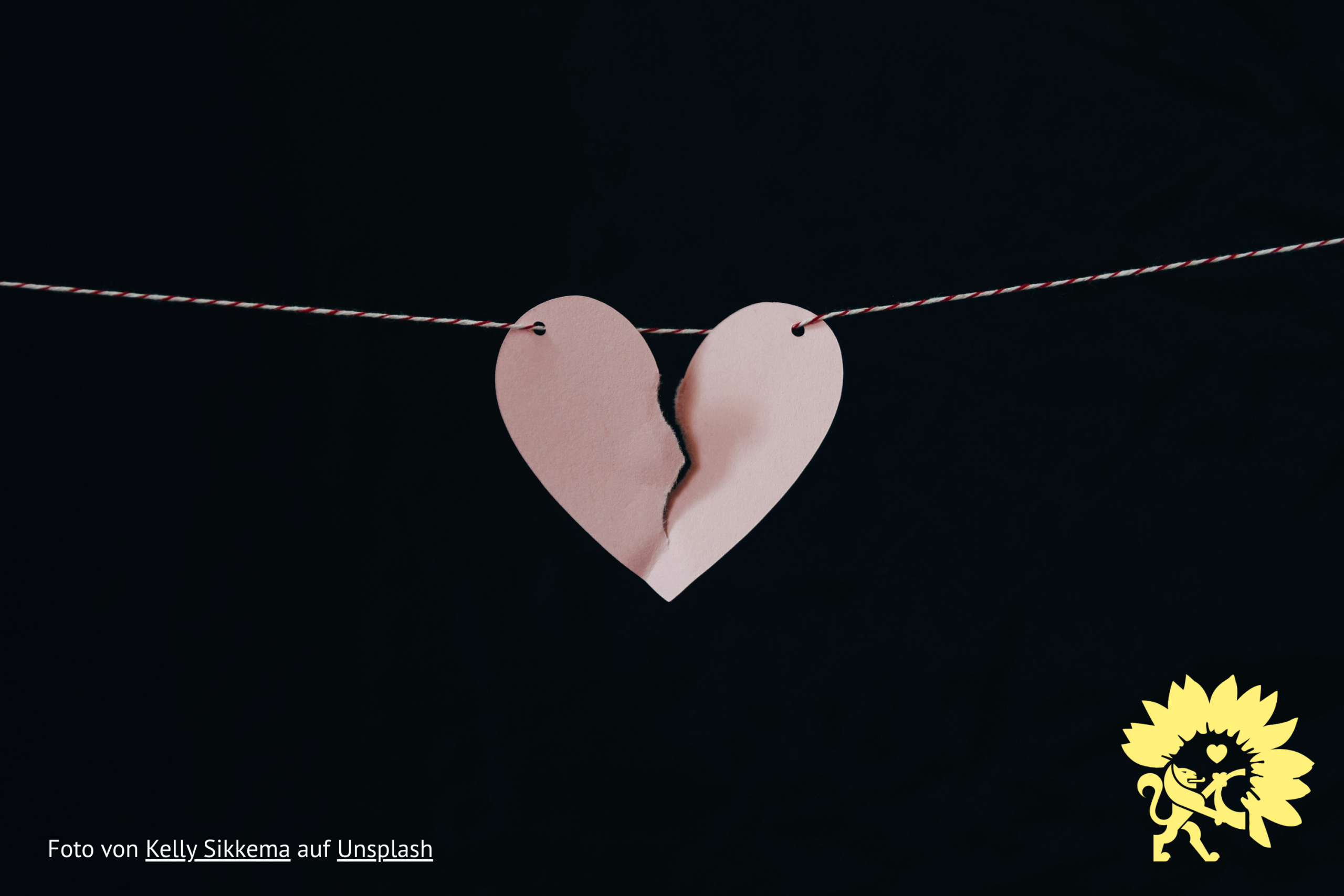 Ein sich spaltendes Herz aus Papier hängt an einer Leine. Unten links steht "Foto von Kelly Sikkema auf Unsplash". Unten rechts die Sonnenblume von BÜNDNIS 90/DIE GRÜNEN mit dem Löwen und einem Herzen aus dem Landkreis-Wappen und -Logo.